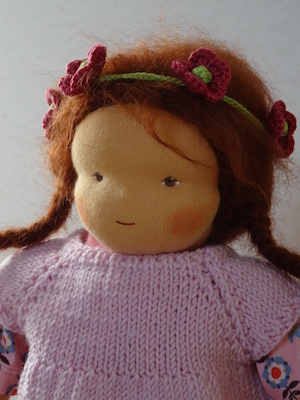 Puppe mit Blumenschmuck in den braunen Haaren und rosa Oberteil. Hat ihren Blick nach links gerichtet
